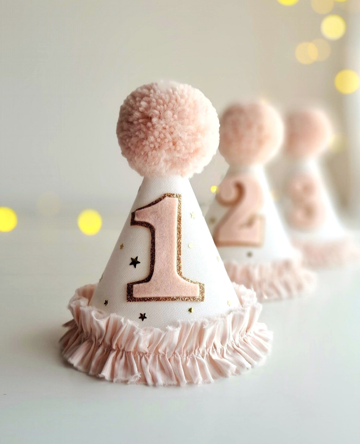 Group of Ivory party hats overlaid with stary gold tulle. The hats have a pink and white stripe ruffle at the bottom and a fluffy pink pompom on the top. By Verity Isabelle.
