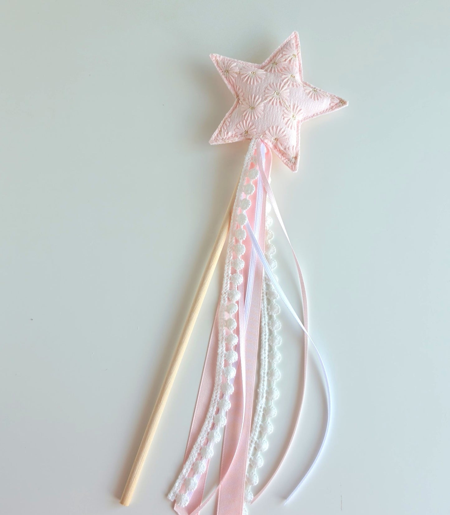 A pink fabric star wand with a wooden handle, decorated with pink and white ribbons, lace trim, set against a plain light gray background.