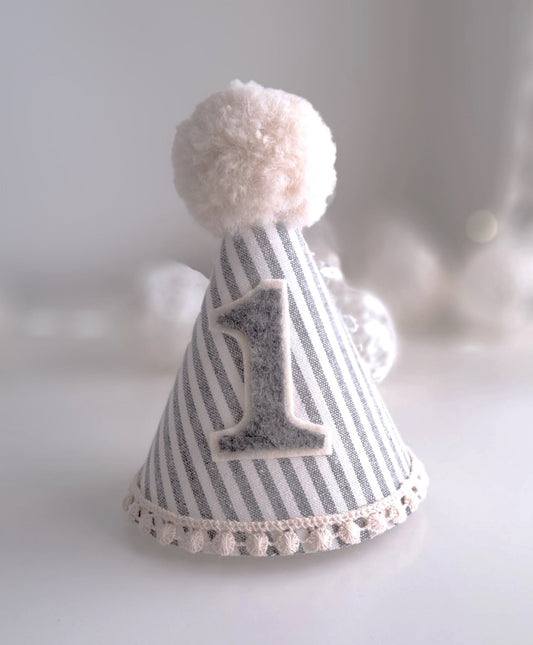 Grey and white pinstripe party hat with a grey felt number one.  The hat has a cream cotton crochet trim with alpaca fluffy pompom on the top.  By Verity Isabelle.
