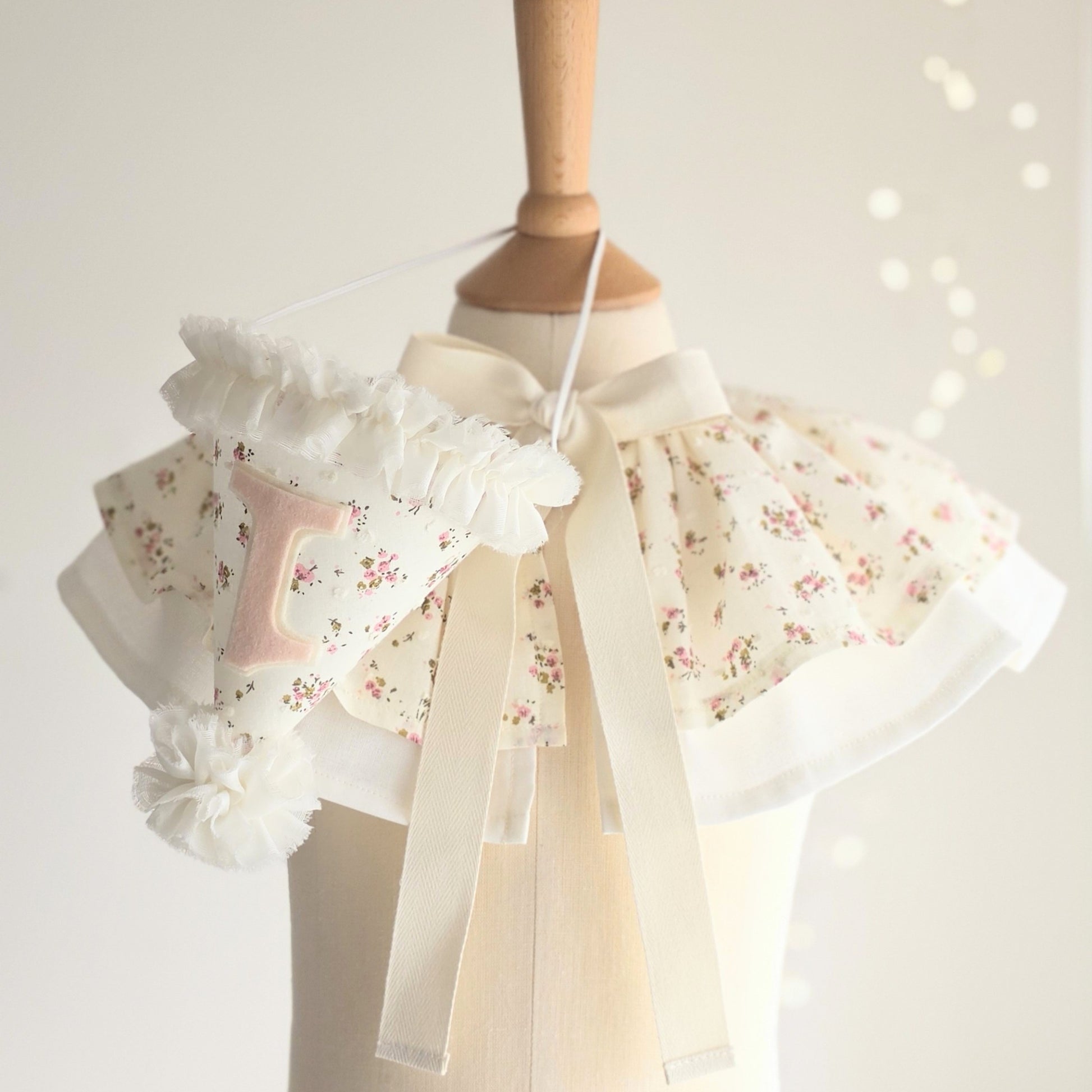 Ditsy floral birthday cape with cream bow ties and matching first birthday party hat displayed on a wooden mannequin.