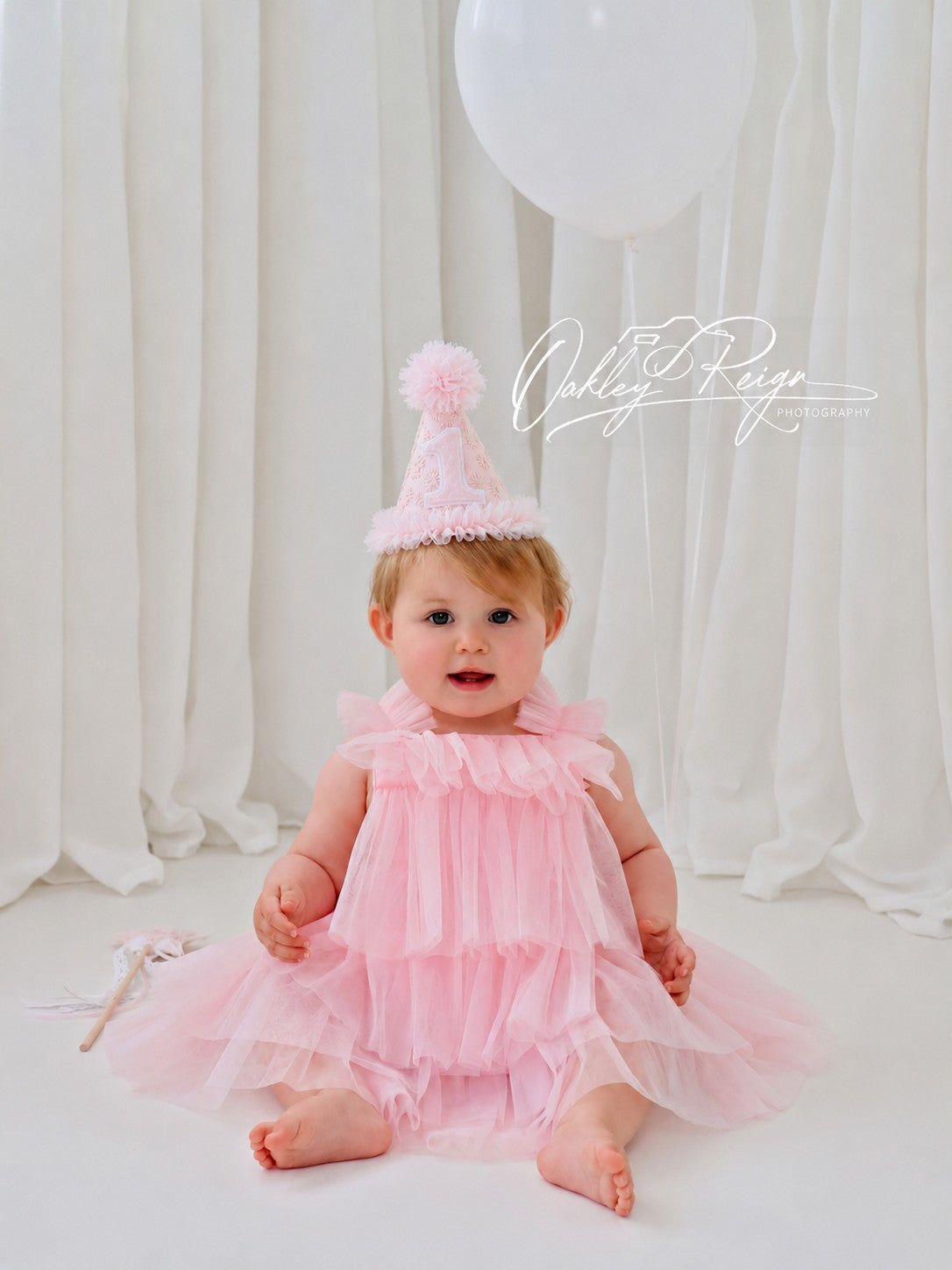 Baby girl sitting in a pale pink tulle dress wearing a Pink Starburst first birthday party hat with chiffon pom-pom and ruffle trim. By Verity Isabelle.