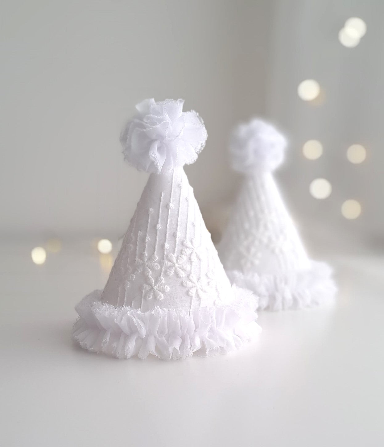 Two white floral lace party hats with chiffon ruffle trims at the bottom and matching pompoms on the top.