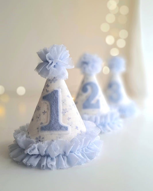 Blue number 1 birthday party hat in front of matching number 2 and 3 hats, with floral fabric and ruffle trim.