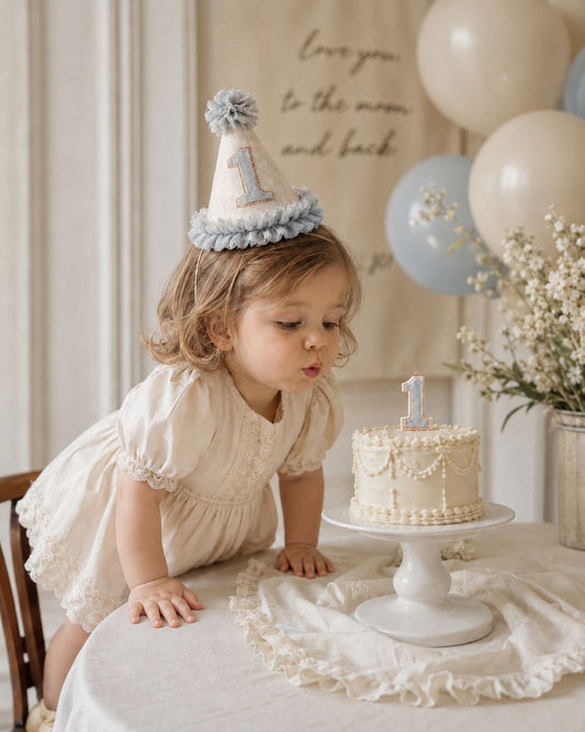 Toddler girl in an ivory dress wearing a dusky blue and cream first birthday party hat, leaning over a table beside a white birthday cake in a soft vintage-style celebration setting.