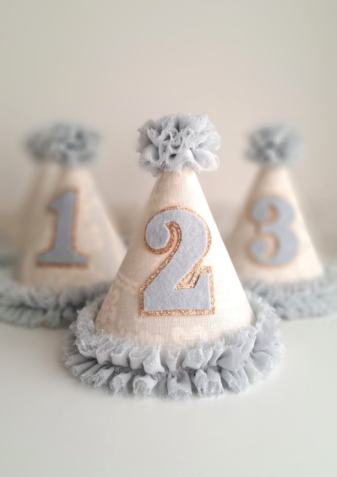 Handmade pale blue number 2 party hat with soft linen fabric, blue ruffled trim and gold stitched detail. Hats with number 1 and number 3 in the background.