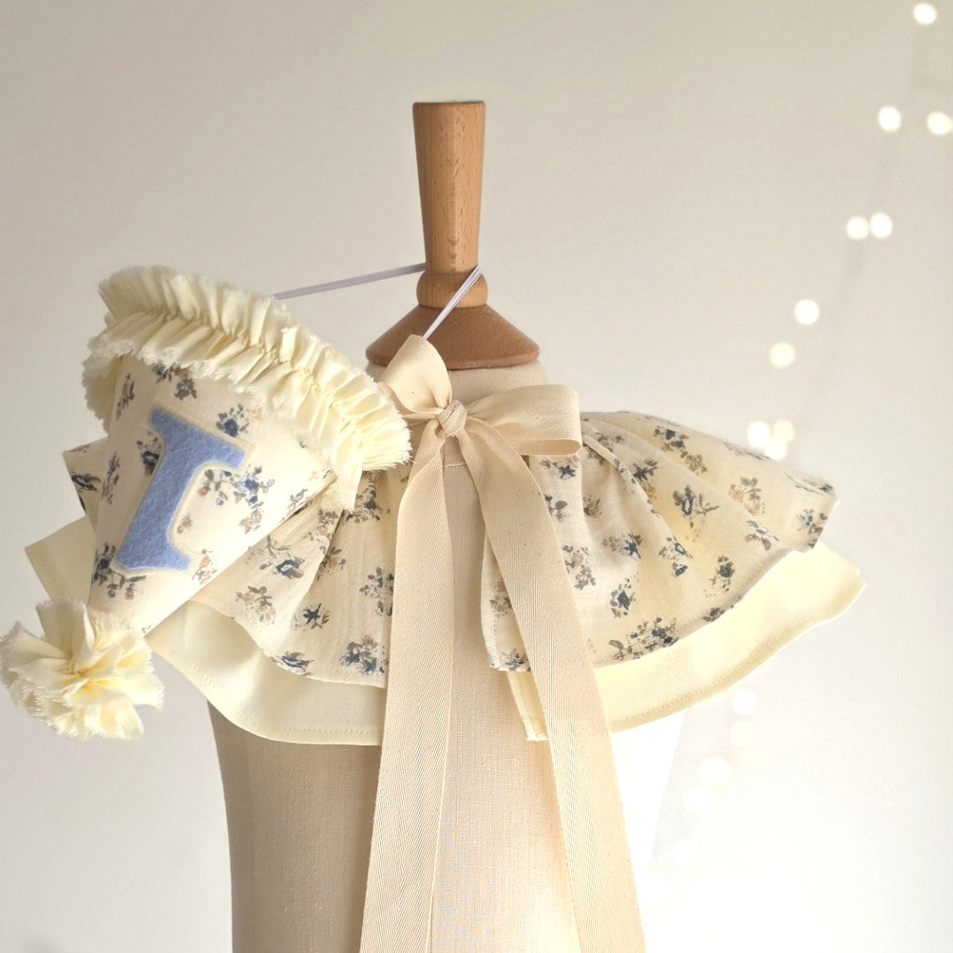 Handmade Mabel floral birthday set featuring a pale lemon floral party hat and matching cape with soft blue ditsy flowers By Verity Isabele. 