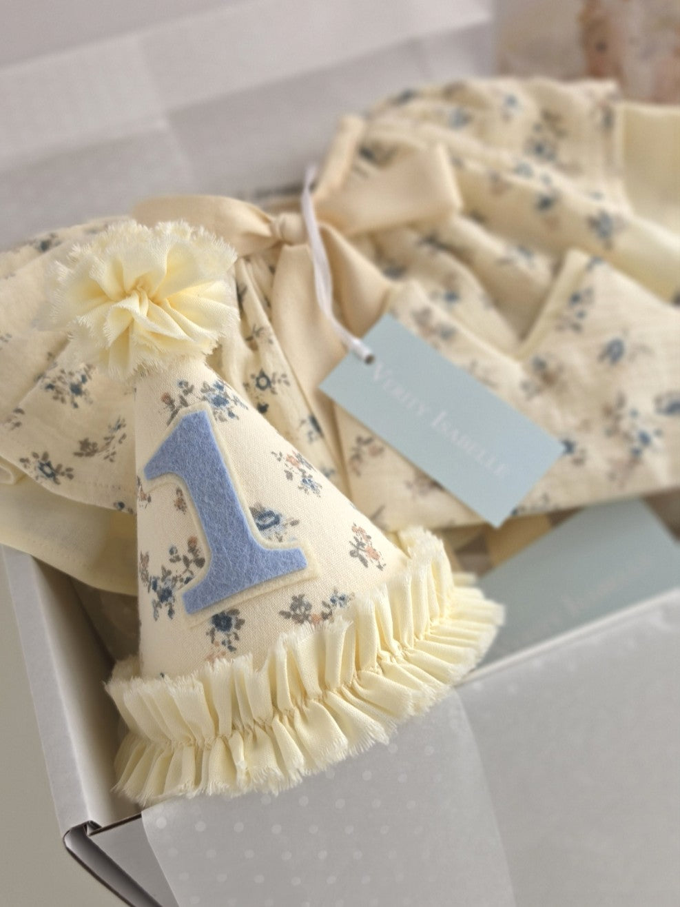 Heirloom-inspired Mabel birthday collection gift box featuring a vanilla floral party hat with blue number one applique, matching cape and soft fabric crown.