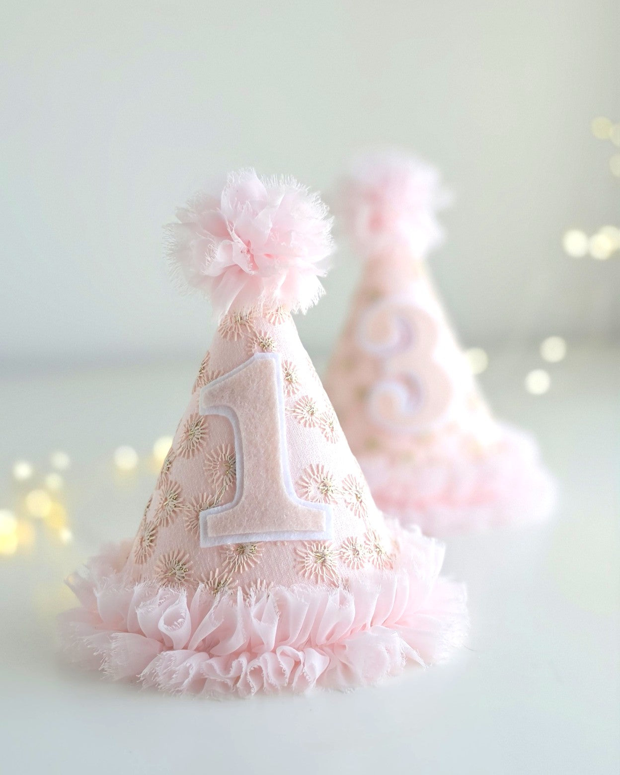Group of Pink Starburst first birthday party hat with felt number 1 and number 3, pale pink embroidered lace, gold starburst detail, chiffon ruffle trim and pom-pom. By Verity Isabelle.