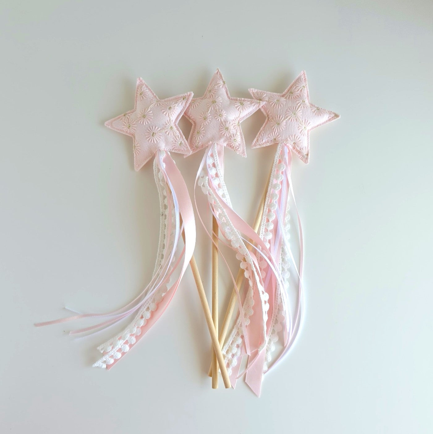 Three pink star wands with embroidered fabric stars, long pink and white ribbons, with a wooden stick handle, arranged on a plain light background.