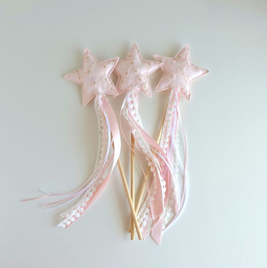 Three pink star wands with embroidered fabric stars, long pink and white ribbons, with a wooden stick handle, arranged on a plain light background.