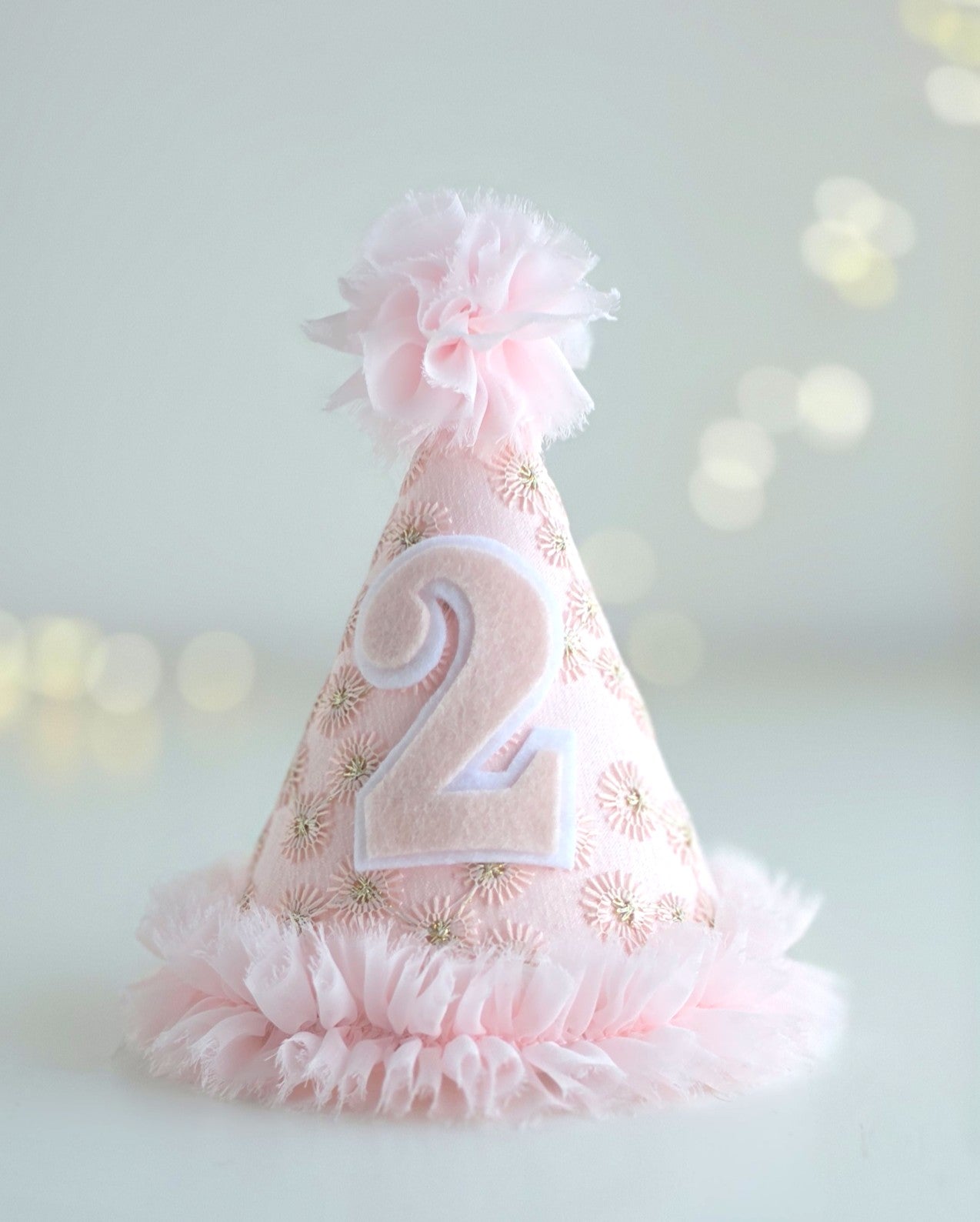 Pink Starburst second birthday party hat with felt number 2, pale pink embroidered lace, gold starburst embroidery, chiffon ruffle trim and pom-pom. By Verity Isabelle.
