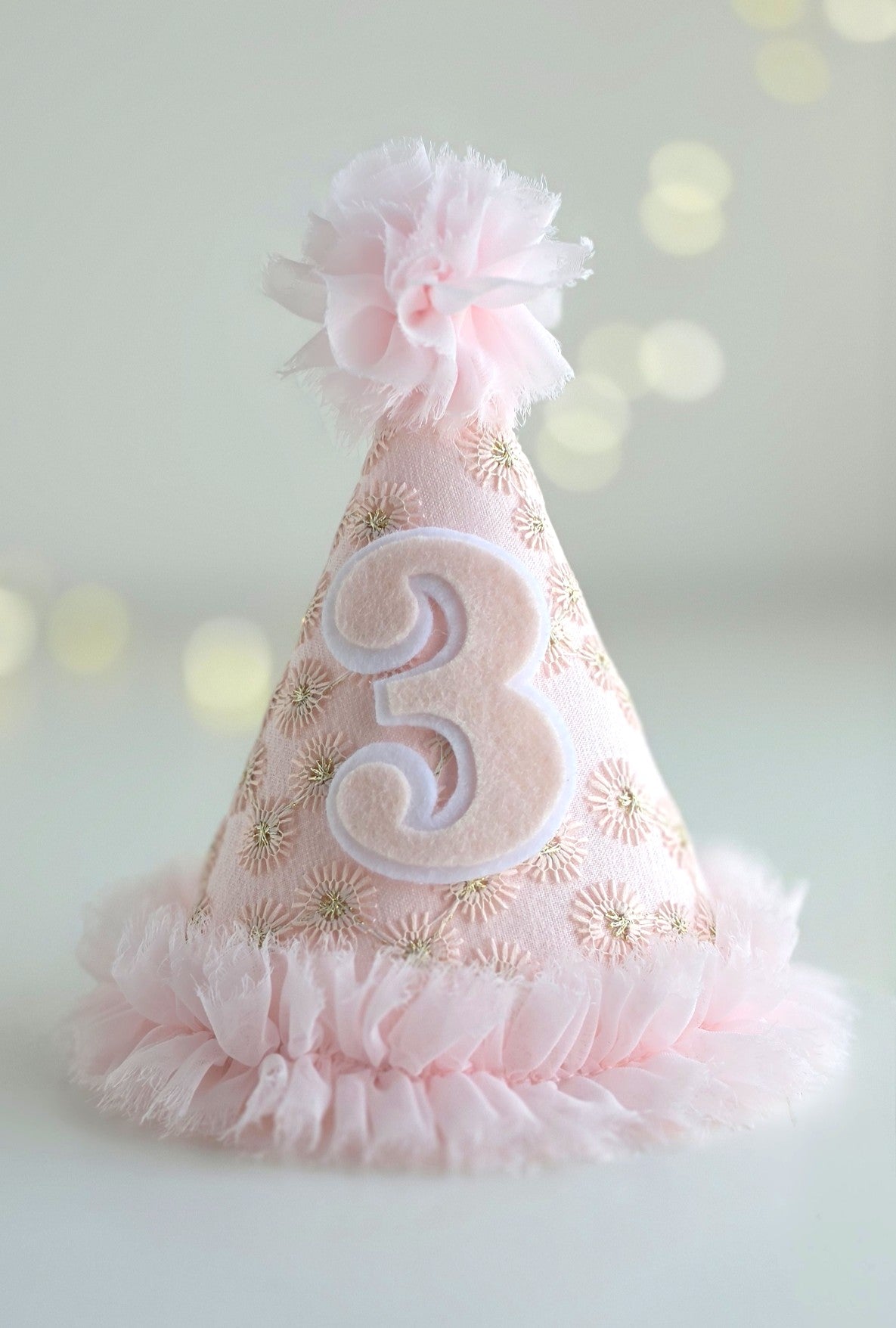 Pink Starburst third birthday party hat with felt number 3, pale pink embroidered lace, gold starburst detail, chiffon ruffle trim and pom-pom. By Verity Isabelle.