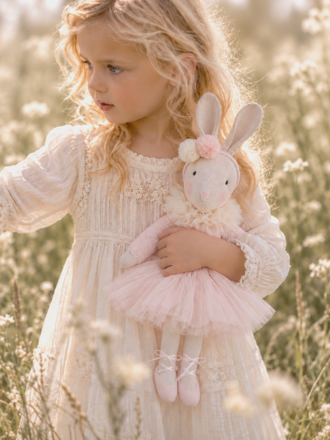 Young girl in a cream dress holding a handmade Verity Ballerina Bunny in a softly lit wildflower meadow.