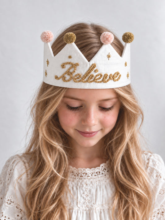 A young girl with long blonde hair wearing a white felt crown with gold glittery Christmas 'Believe' lettering, gold star details, and pink and gold pom-poms, set against a light background.