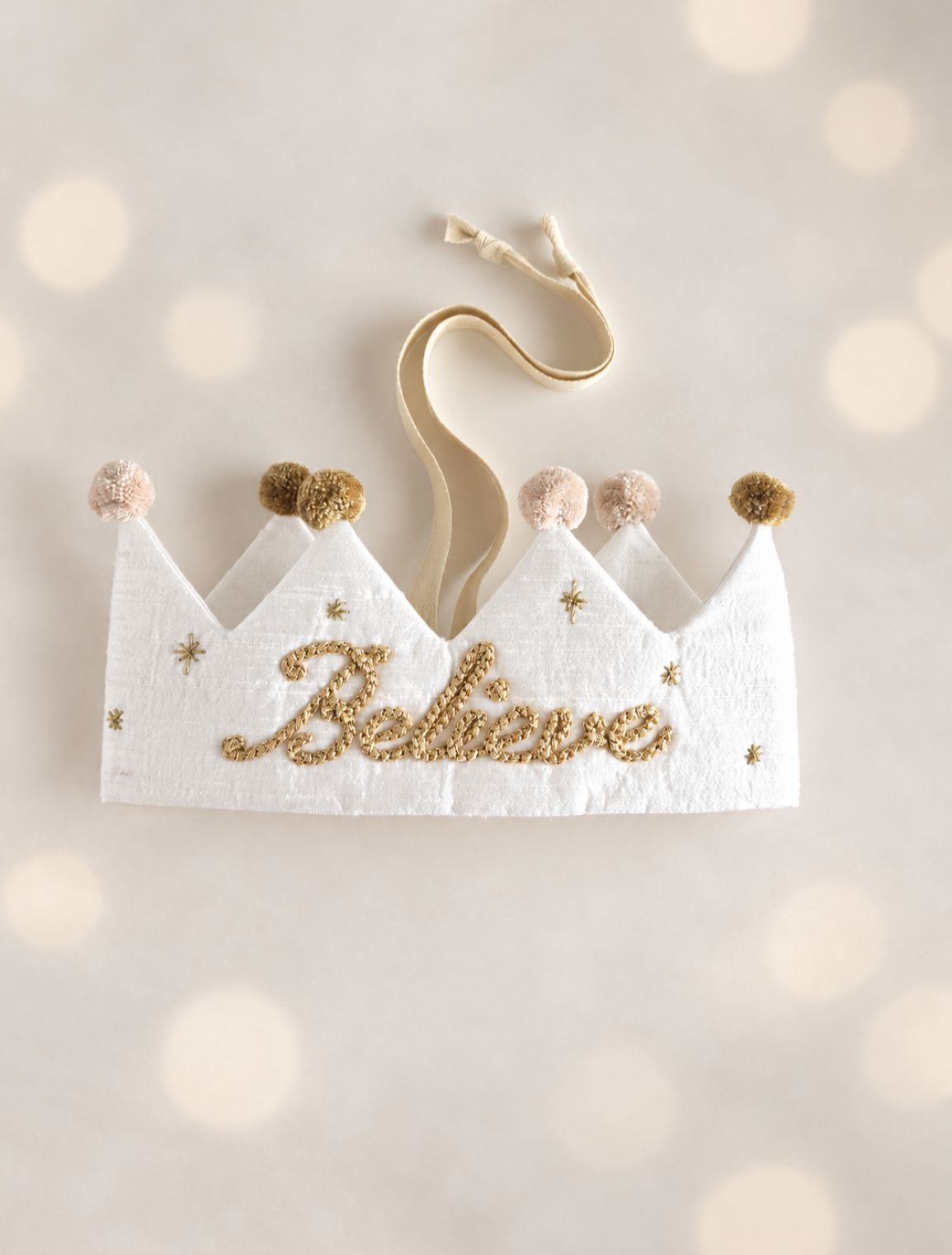 Believe silk Christmas crown laid flat showing adjustable ribbon ties, gold embroidery and cotton pom-poms.