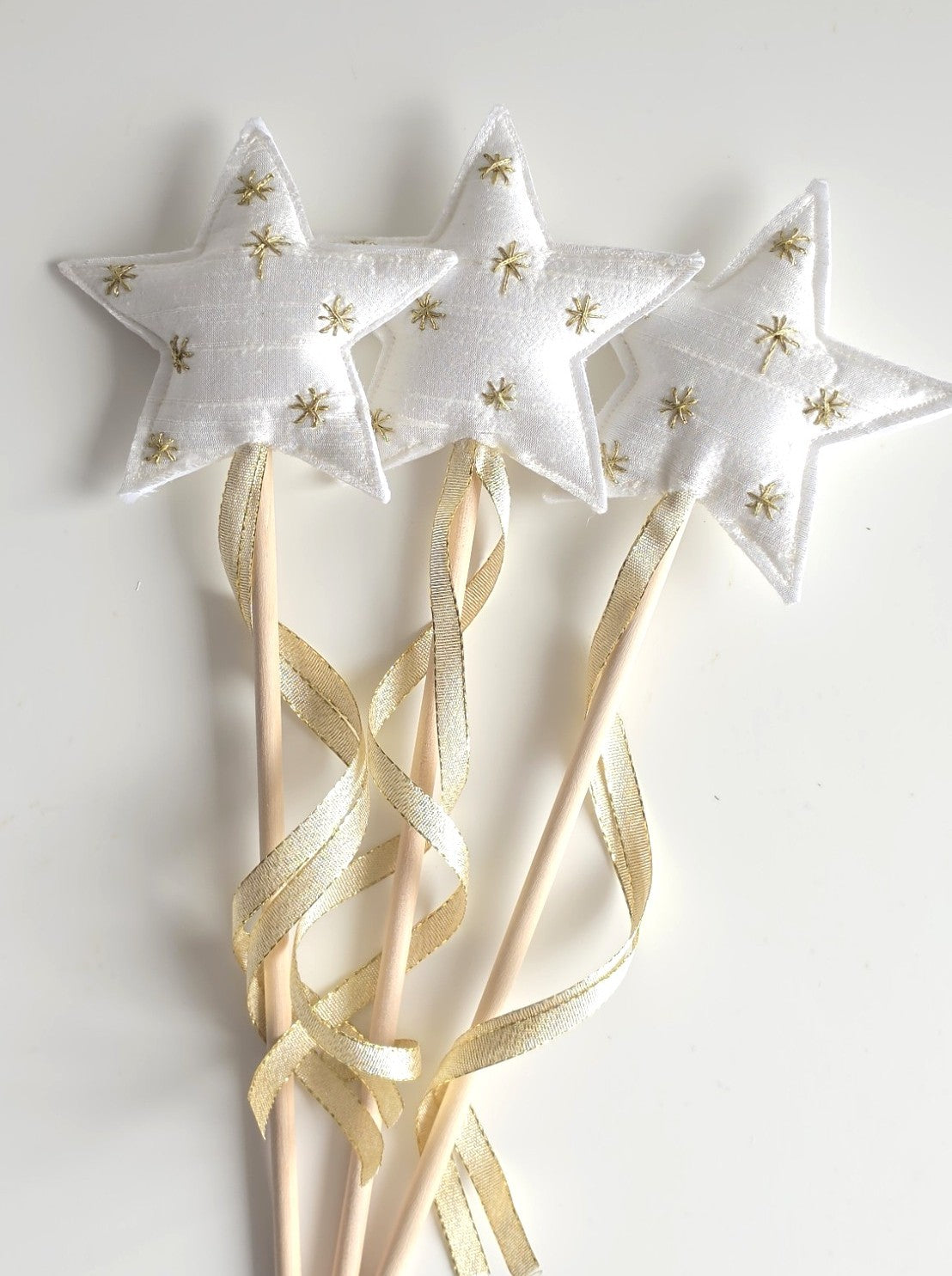 Three white satin star-shaped wand toppers with gold embroidered stars, attached to wooden sticks with cream ribbons, on a white background.