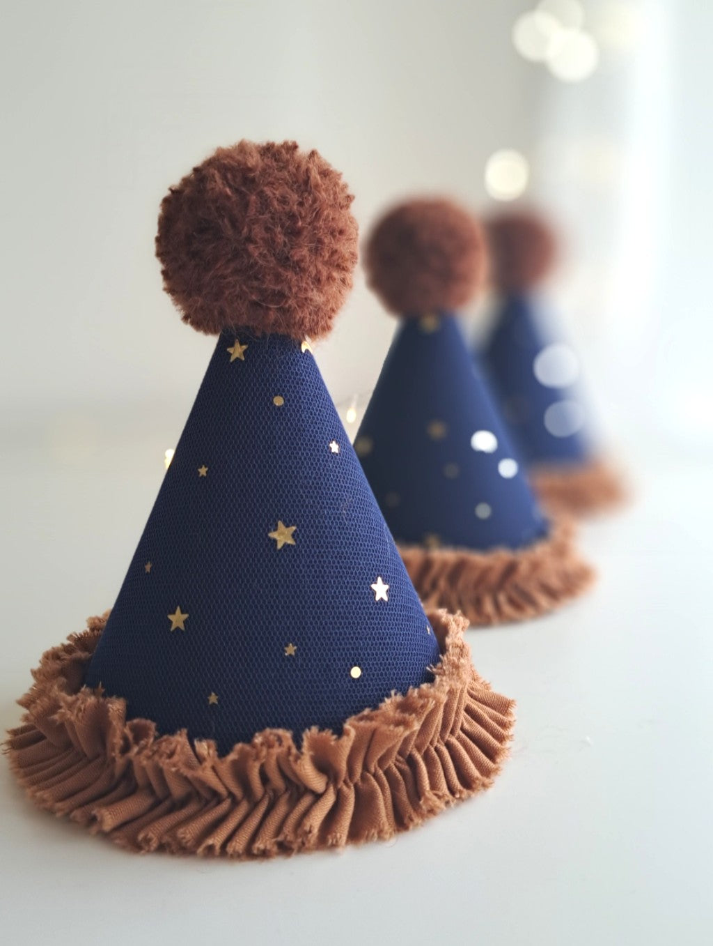 A row of midnight navy linen party hats with gold star tulle, ginger fabric frill, and alpaca pom-poms — handmade for imaginative play or seasonal dressing-up.
